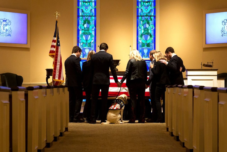 A group of people dressed in black gathered around a coffin draped in an US flag, with a yellow labrador sitting beside it.