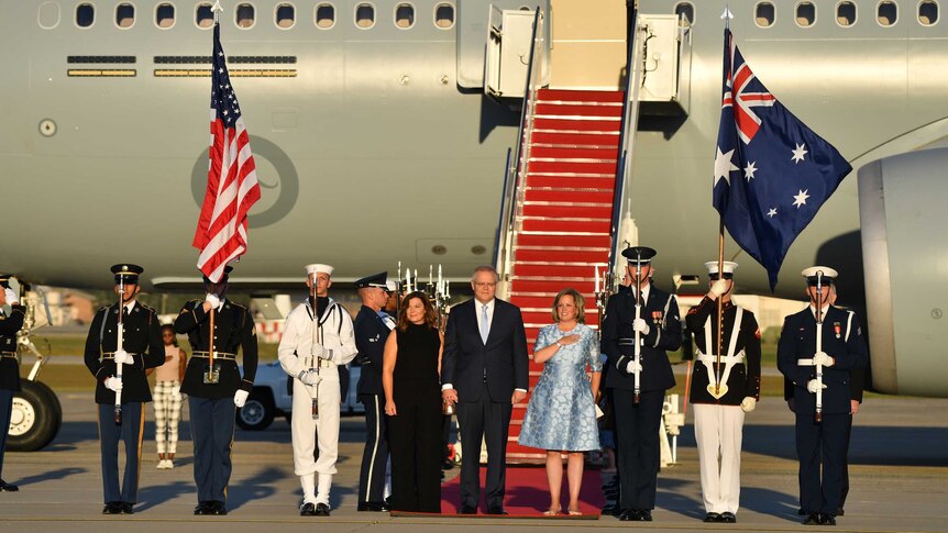 Scott and Jenny Morrison stand with US military personnel at the bottom of a set of aeroplane stairs.