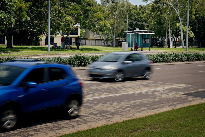 Two cars drive along a stretch of road. A bus stop is in the background