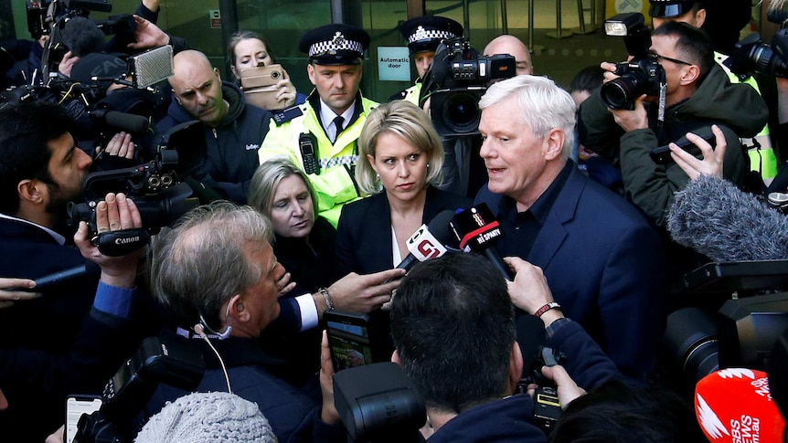 A throng of journalists gather around two people in suits while two British police officers wear hi-vis behind them.