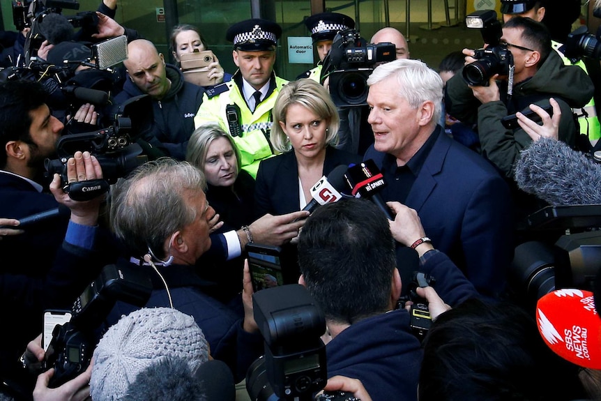 A throng of journalists gather around two people in suits while two British police officers wear hi-vis behind them.