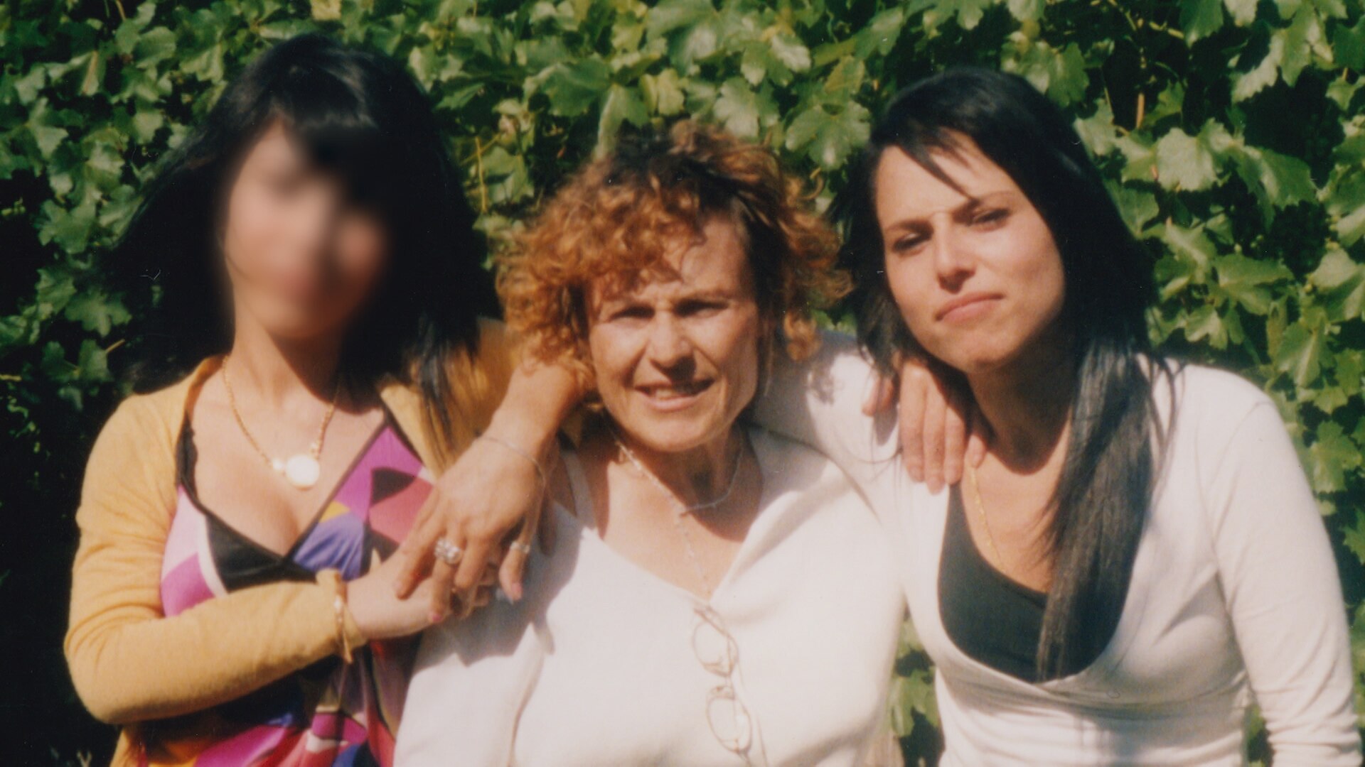 A woman stands in front of shrubbery with her two adult daughters, smiling. One daughter's face blurred
