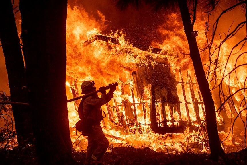 A firefighter in protective gear holds a thick hose over his right shoulder and aims it at a destroyed house still ablaze