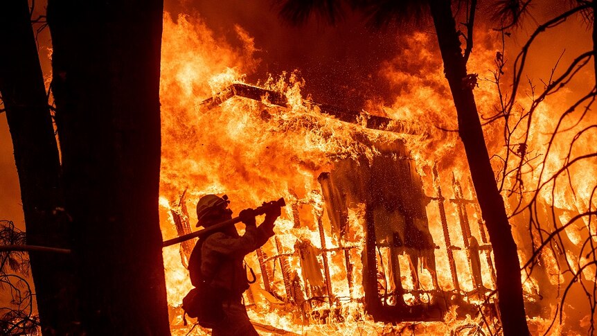 A firefighter in protective gear holds a thick hose over his right shoulder and aims it at a destroyed house still ablaze