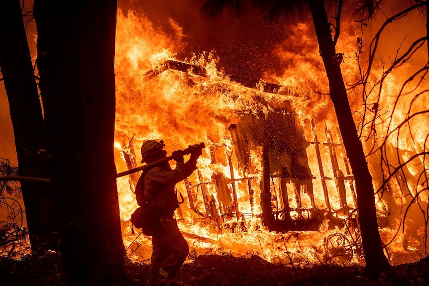 A firefighter in protective gear holds a thick hose over his right shoulder and aims it at a destroyed house still ablaze