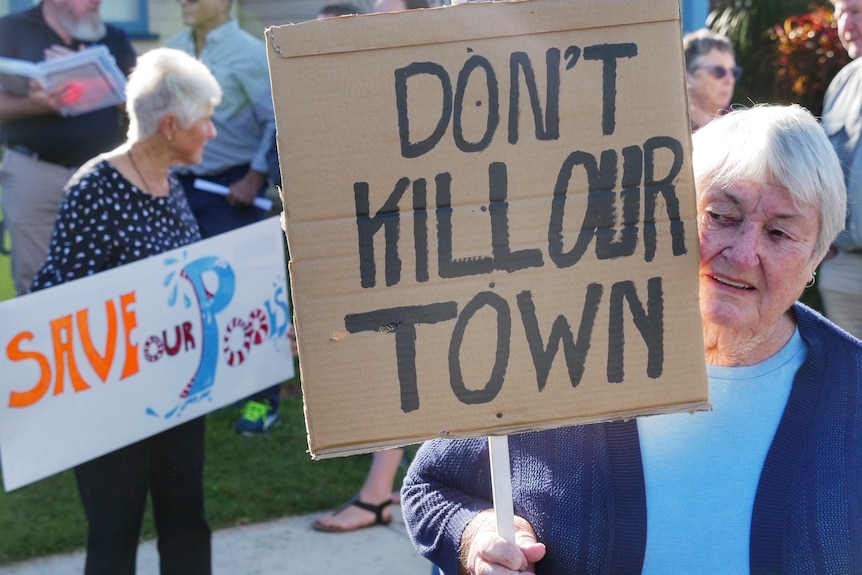 An elderly, white-haired woman amid a crowd of protesters holds a cardboard sign that reads "don't kill our town".