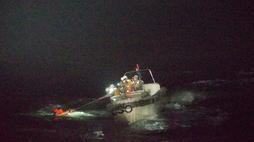 At night, a man in a life jacket is pulled towards a small boat by a line.