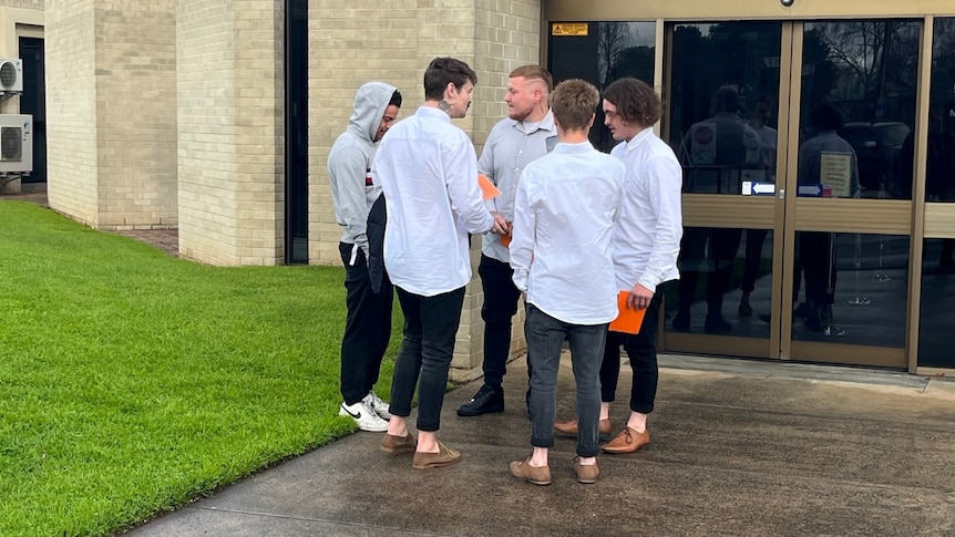 Four young men standing outside a court building, all wear white shirts, black jeans, one wears grey hoodie, grass on left.