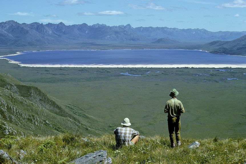 Two hikers look out at a lake, with mountains in the background.