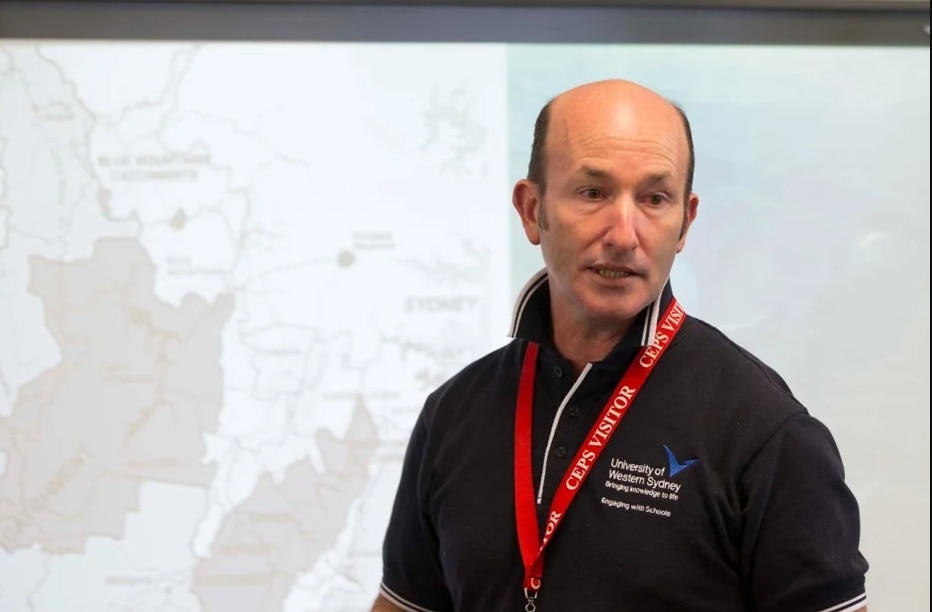 a man in a black polo shirt giving a presentation at a university