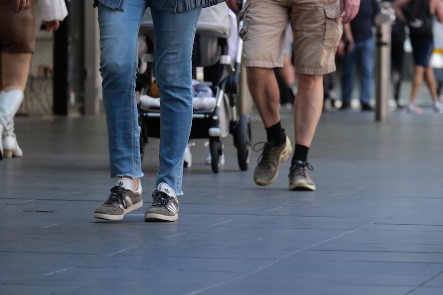 Two pairs of feet and lower legs in focus walking on pavement with a crowd blurred behind.