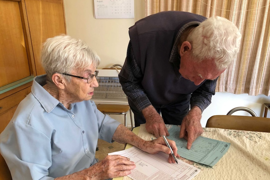 A lady wearing a blue shirt and a man wearing a blue jumper are sitting at a table writing with a pen on a piece of paper