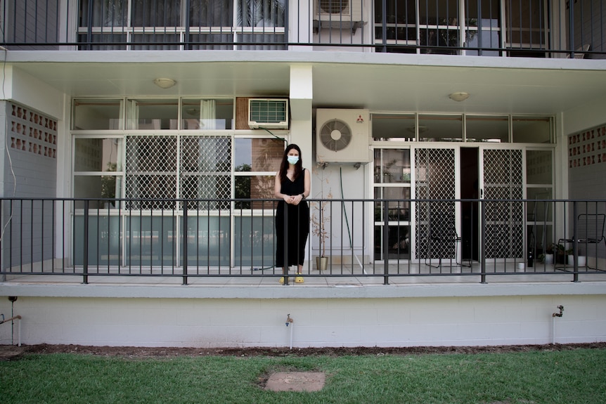 A woman wearing a surgical mask standing on the balcony of an apartment.