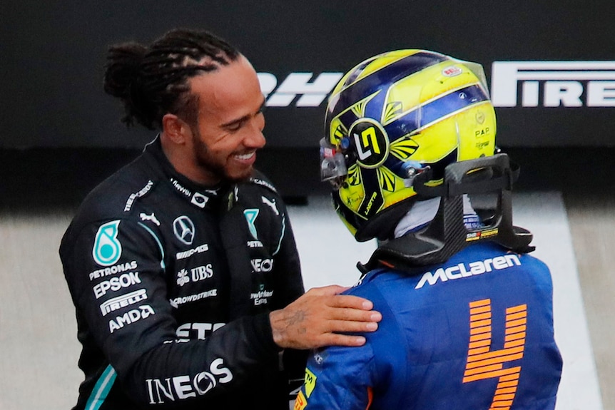 Lewis Hamilton smiles as he greets a helmeted man in yellow and blue near a race track.