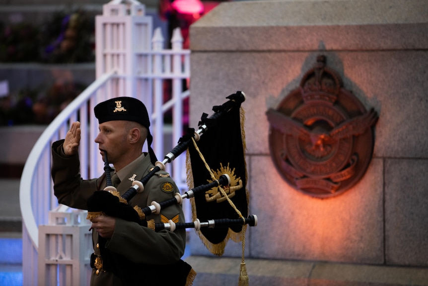 A man salutes while holding bag pipes in front of the memorial