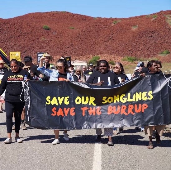Protestors walk and hold a sign reading 'save our songlines save the burrup'