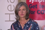 A woman with grey hair and a blue shirt standing in front of a banner