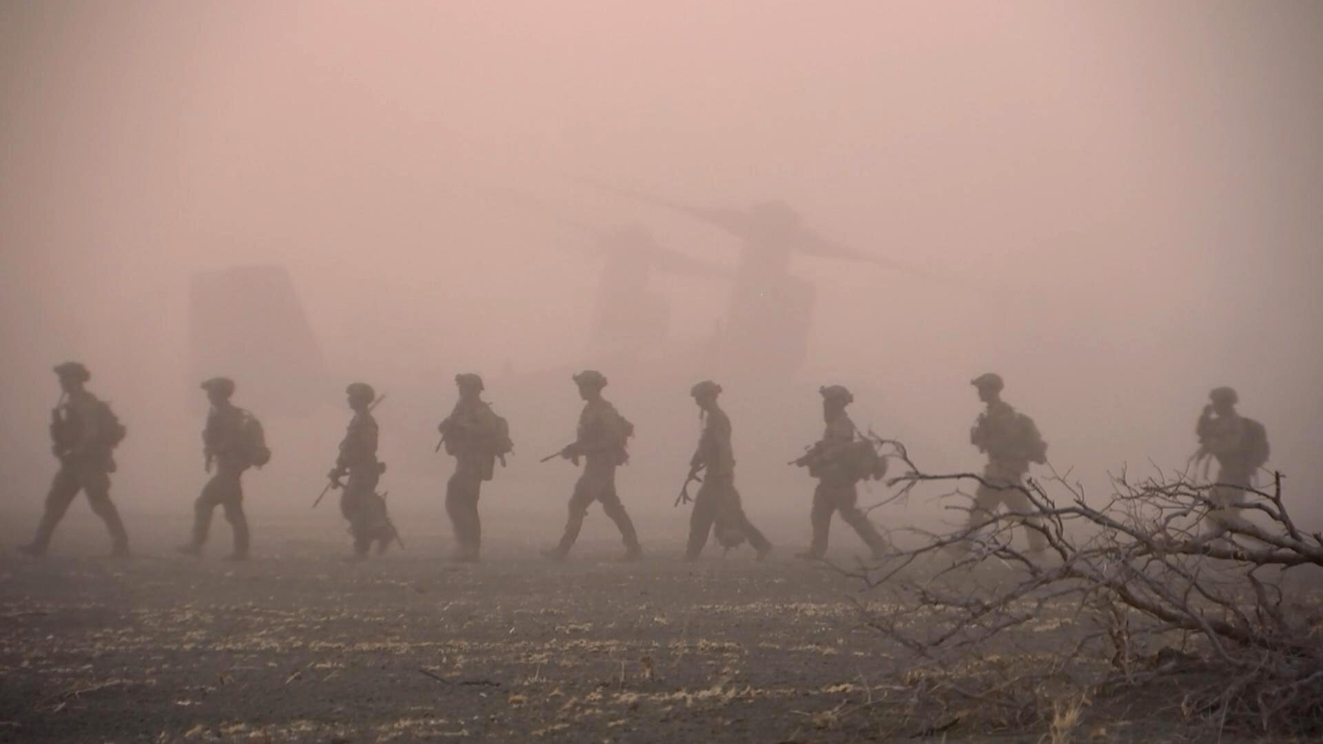 A line of soldiers silhouetted in the dust from a helicopter, march in single file. They are wearing gear and holding rifles.