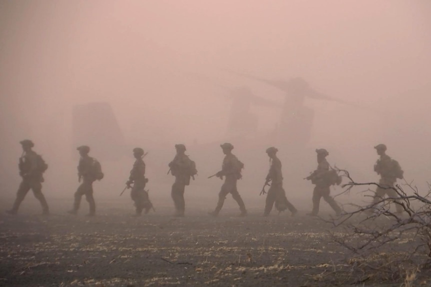 A line of soldiers silhouetted in the dust from a helicopter, march in single file. They are wearing gear and holding rifles.