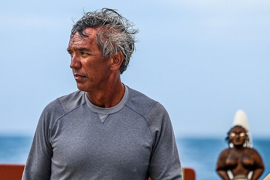 Hawaiian man in grey shirt looks to his right shoulder with blue ocean in background. 