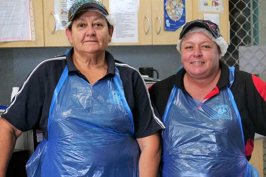 Robyn Greenwood and Vanessa Hancock wearing plastic aprons, hair nets and caps in a kitchen, cupboards in background.
