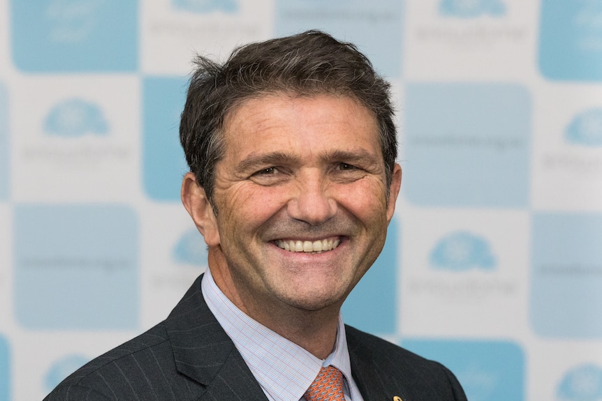 Headshot of a smiling middle aged caucasian man, black suit, orange pattern tie, blue shirt, stands against blue and white logo.