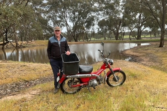 a man and a small motorcycle stand in front of a dam in a field