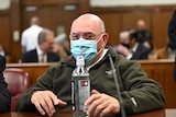 A man is sitting at a desk in a court room wearing a green sweater and blue surgical mask. 