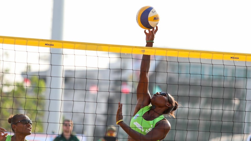 Female volleyball player in green top reaches for ball behind net.