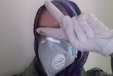 A woman pictured gesturing the peace sign wearing hijab, gloves and a face mask.