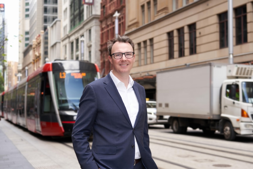 Dean wears a blue suit and white shirt and smiles as a tram approaches behind him.