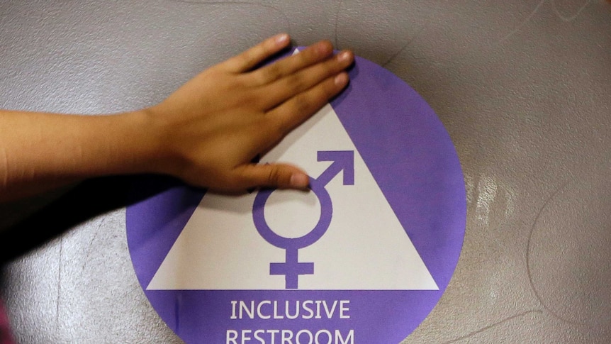 A hand rests on an "inclusive restroom" sign on a toilet door