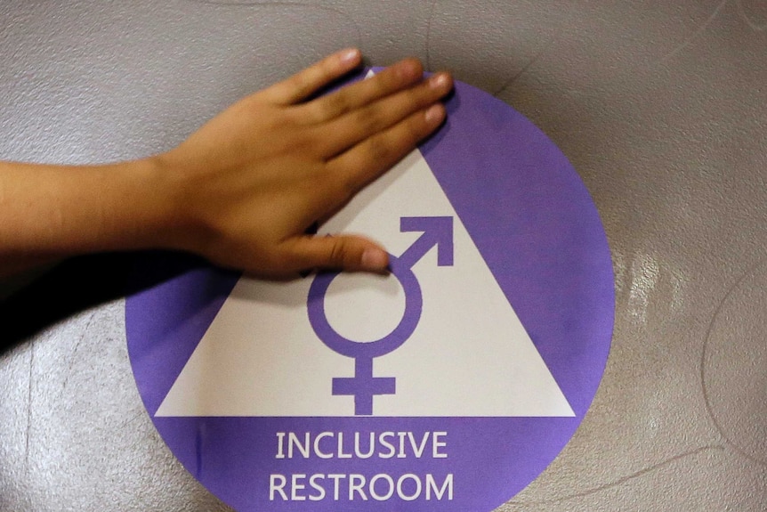 A hand rests on an "inclusive restroom" sign on a toilet door