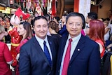 Two men wearing suits stand in busy street with many people behind them