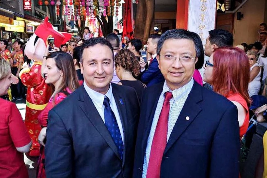 Two men wearing suits stand in busy street with many people behind them