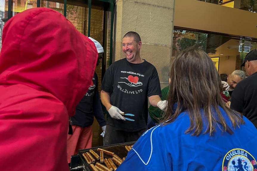 A man holding a pair of tongs smiles as he and stands next to a barbeque with other people around him.