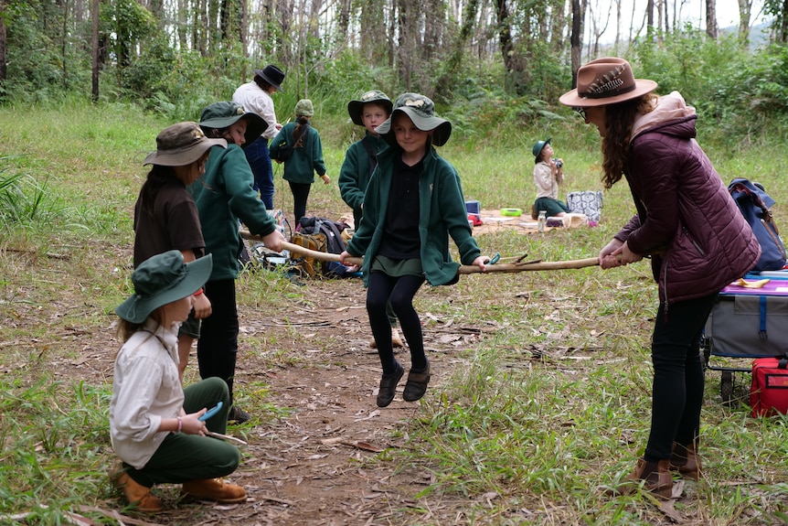 The school's principal helps a student hold up a long stick for another student to sit on to learn about balance