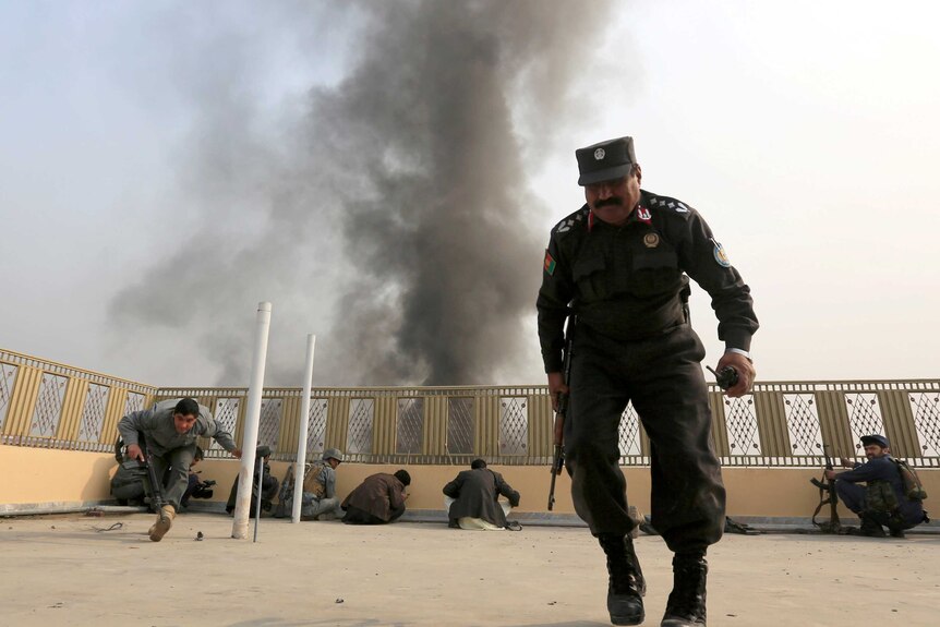 Afghan police officers take position hide behind the fence on the rooftop of a building as smoke fills the sky.