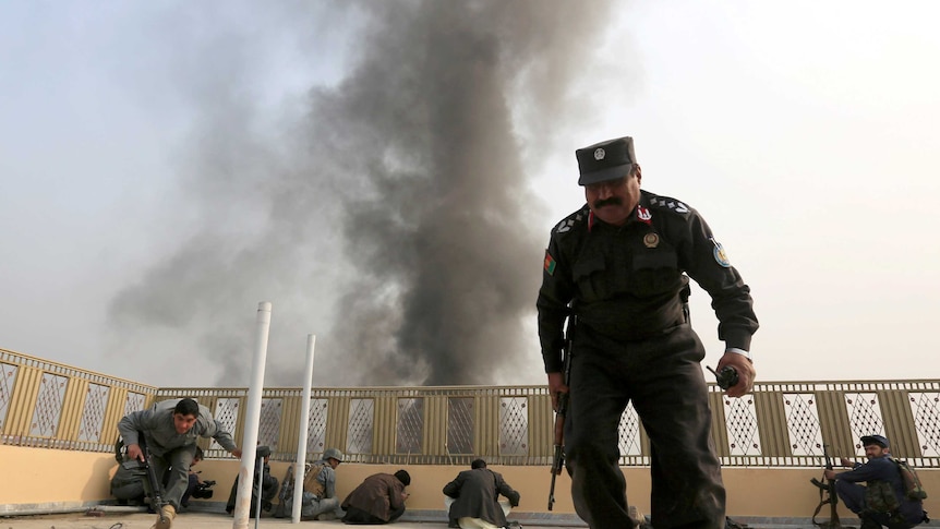 Afghan police officers take position hide behind the fence on the rooftop of a building as smoke fills the sky.
