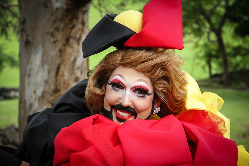 A smiling drag queen wears a bright hat and puffy neck colour in red, yellow and blackn