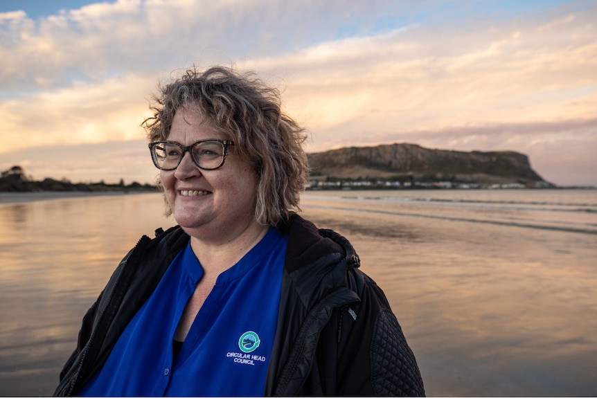 A woman with glasses stands on a beach in front of a promontory