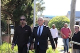 A bald man in a suit walking near a carpark to court, standing next to a man in black clothing with a pixelated face.