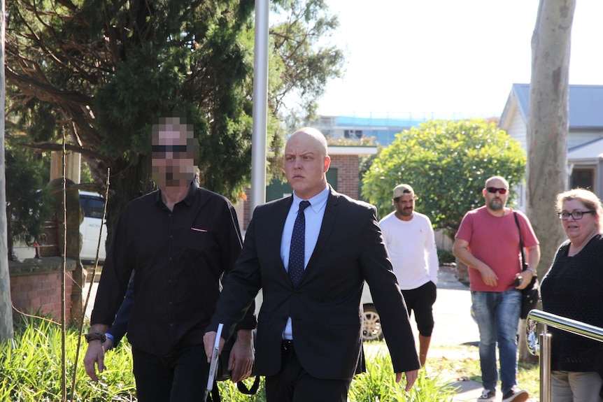A bald man in a suit walking near a carpark to court, standing next to a man in black clothing with a pixelated face.