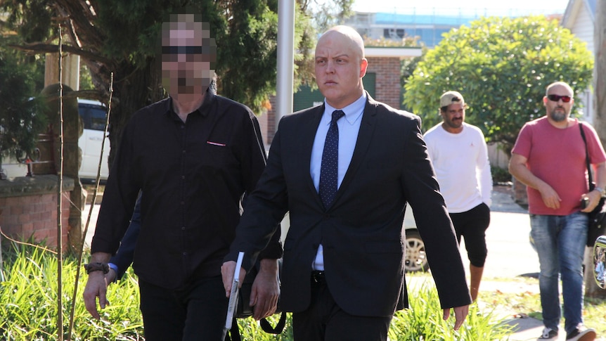 A bald man in a suit walking near a carpark to court, standing next to a man in black clothing with a pixelated face.