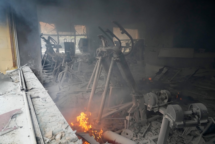 Excercise bikes are shown in a room, destroyed and covered in soot, with flames visible