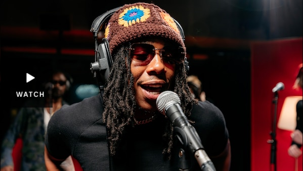 Man wearing a brown beanie with yellow flowers, sunglasses singing into a microphone in a studio. Has Video.