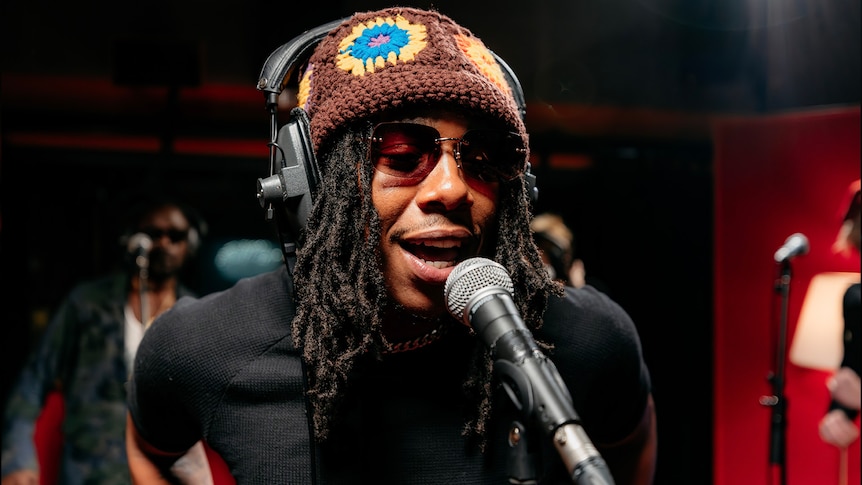 Man wearing a brown beanie with yellow flowers, sunglasses singing into a microphone in a studio