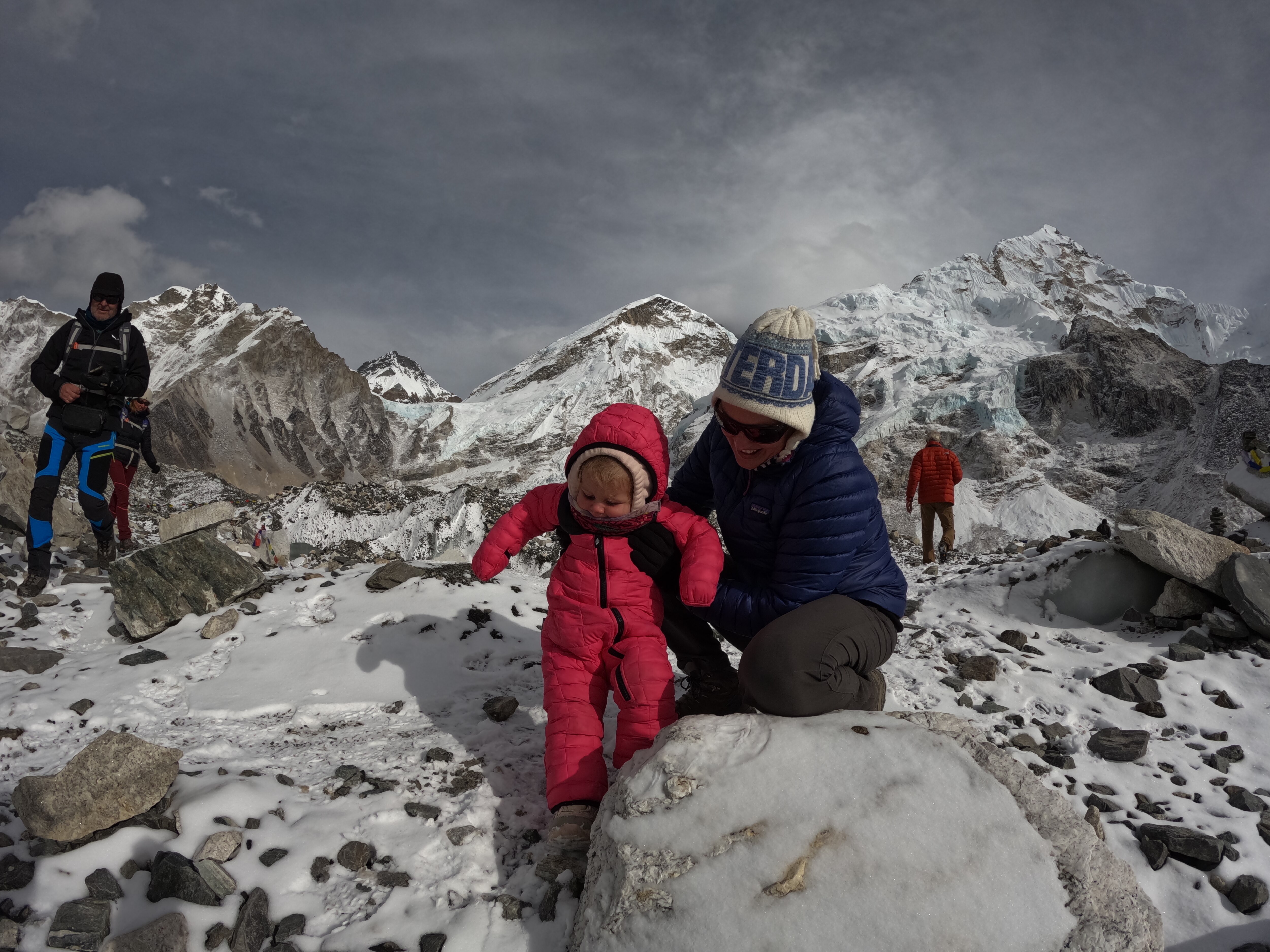 A  smiling woman holds her little daughter in a snowsuit as her squat on snowy mountains.