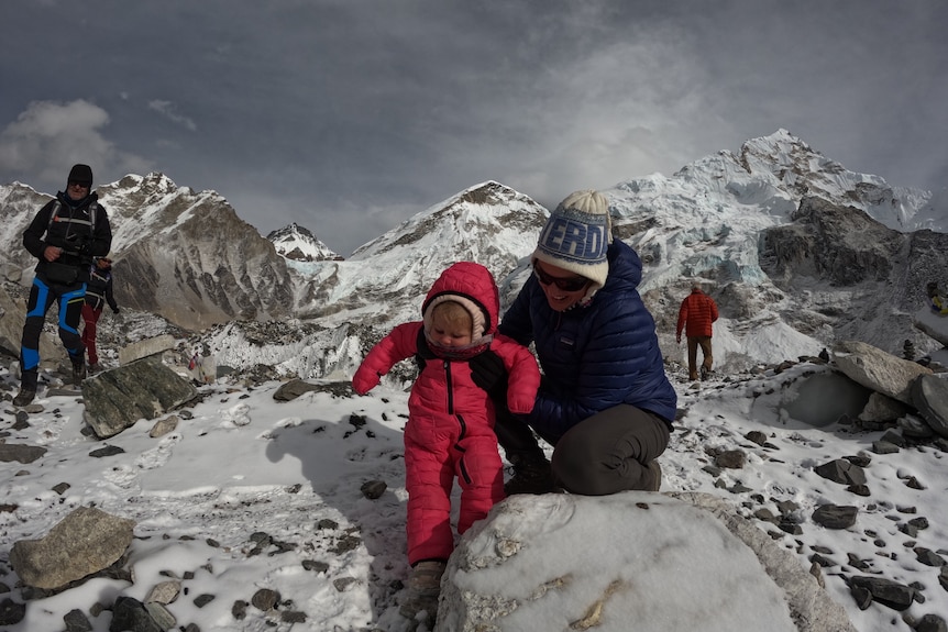 A  smiling woman holds her little daughter in a snowsuit as her squat on snowy mountains.