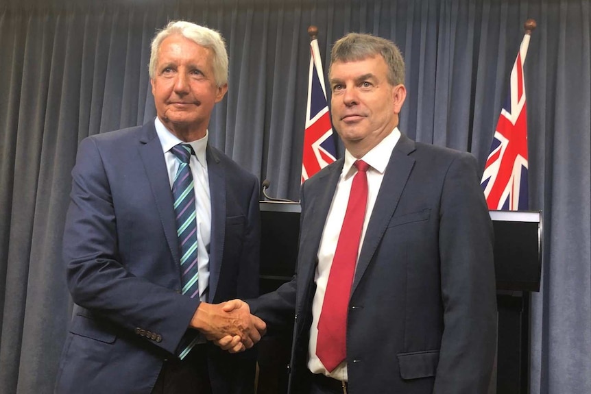 Two men in suits shake hands in front of a dais with flags in the background.